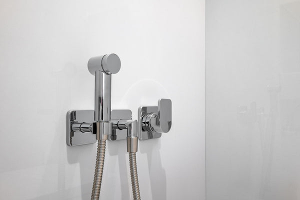 Chrome handheld bidet sprayer with flexible hose and wall-mounted control valve on a white bathroom wall.