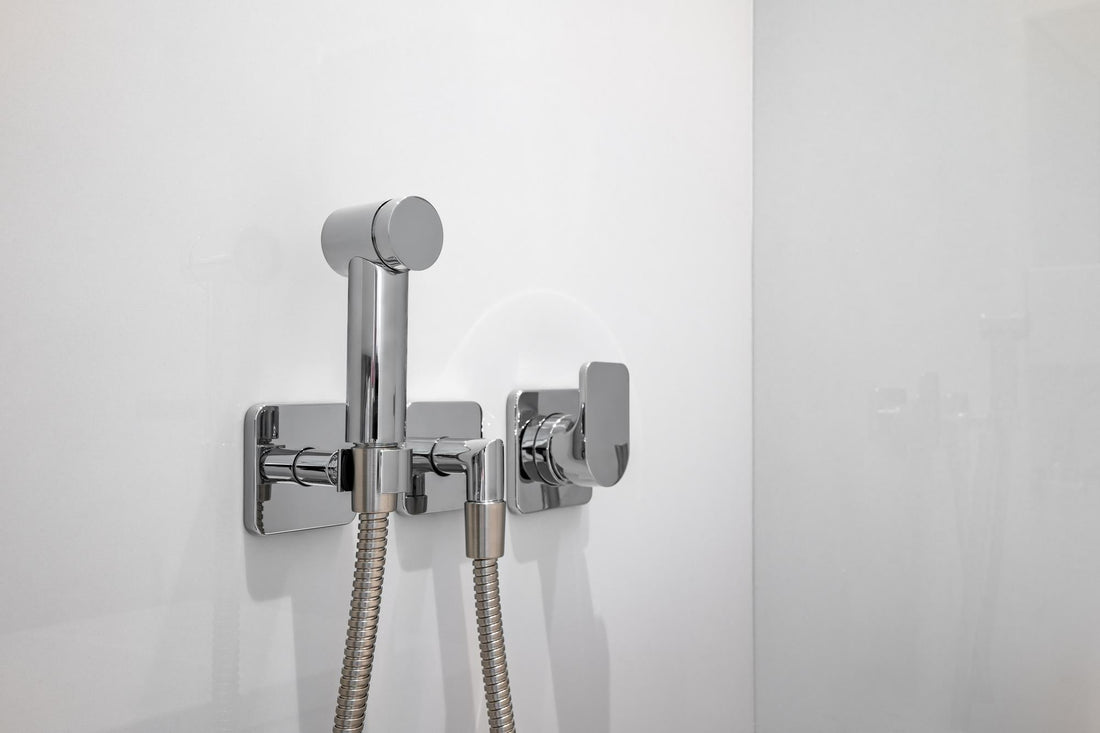 Chrome handheld bidet sprayer with flexible hose and wall-mounted control valve on a white bathroom wall.