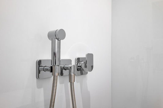 Chrome handheld bidet sprayer with flexible hose and wall-mounted control valve on a white bathroom wall.